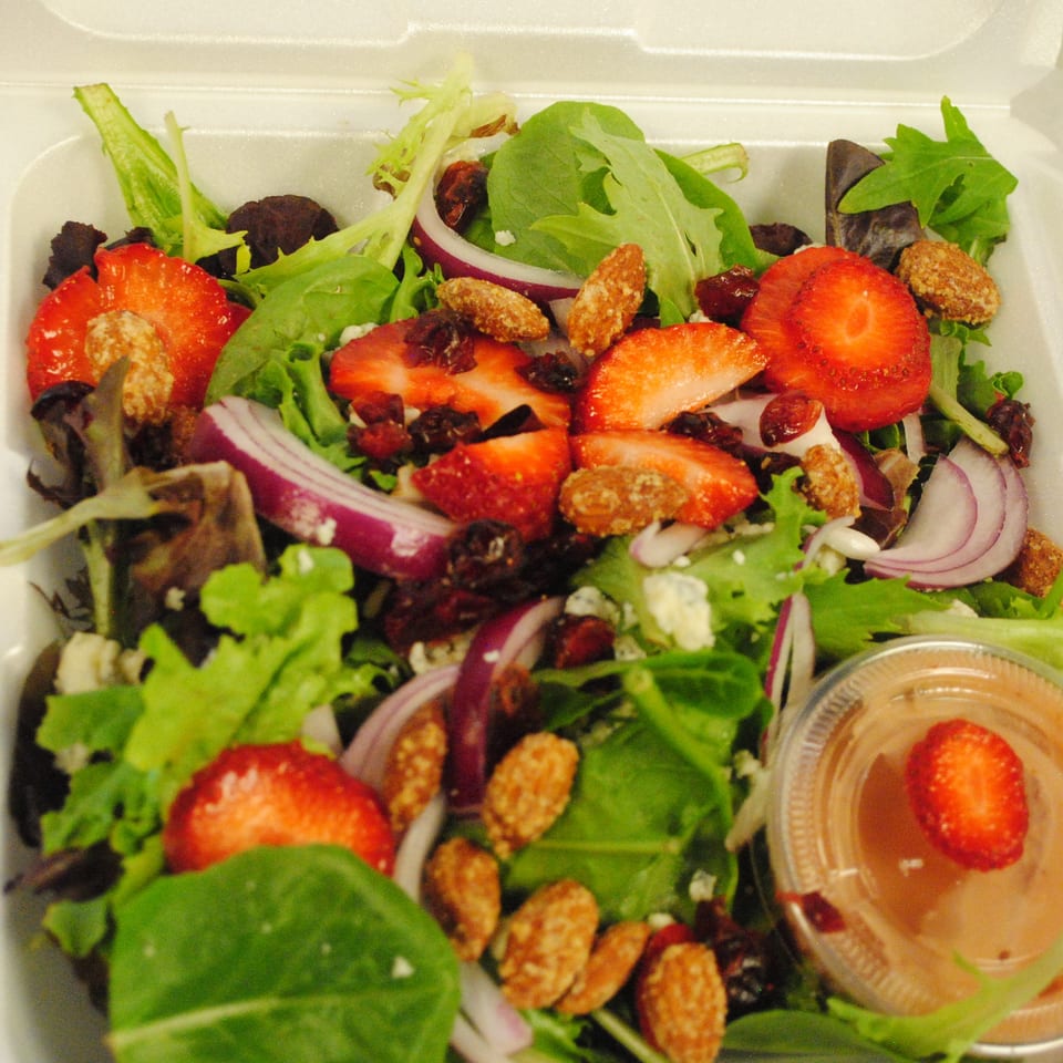 A fresh salad with strawberries, red onions, candied nuts, and a side of dressing in a takeout container.