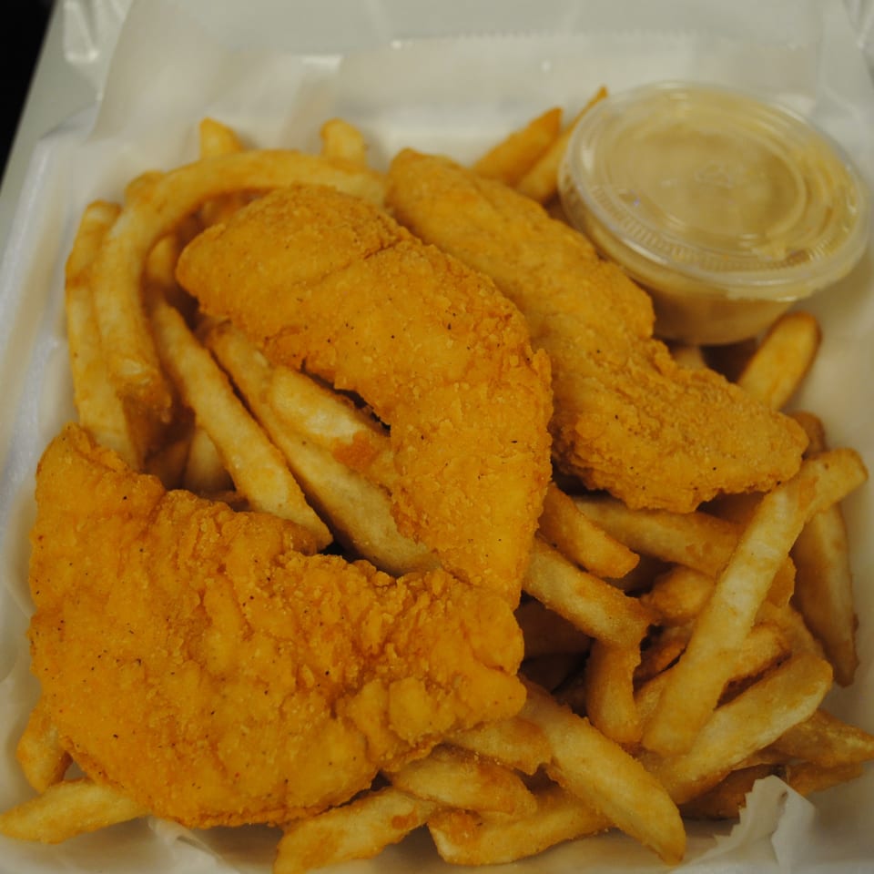 Golden-brown fried chicken tenders and french fries in a white takeout container with a side of sauce.